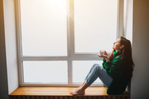 Woman sitting by a window with warm morning light, taking a quiet moment to reset during a winter energy slump and support natural wellness.